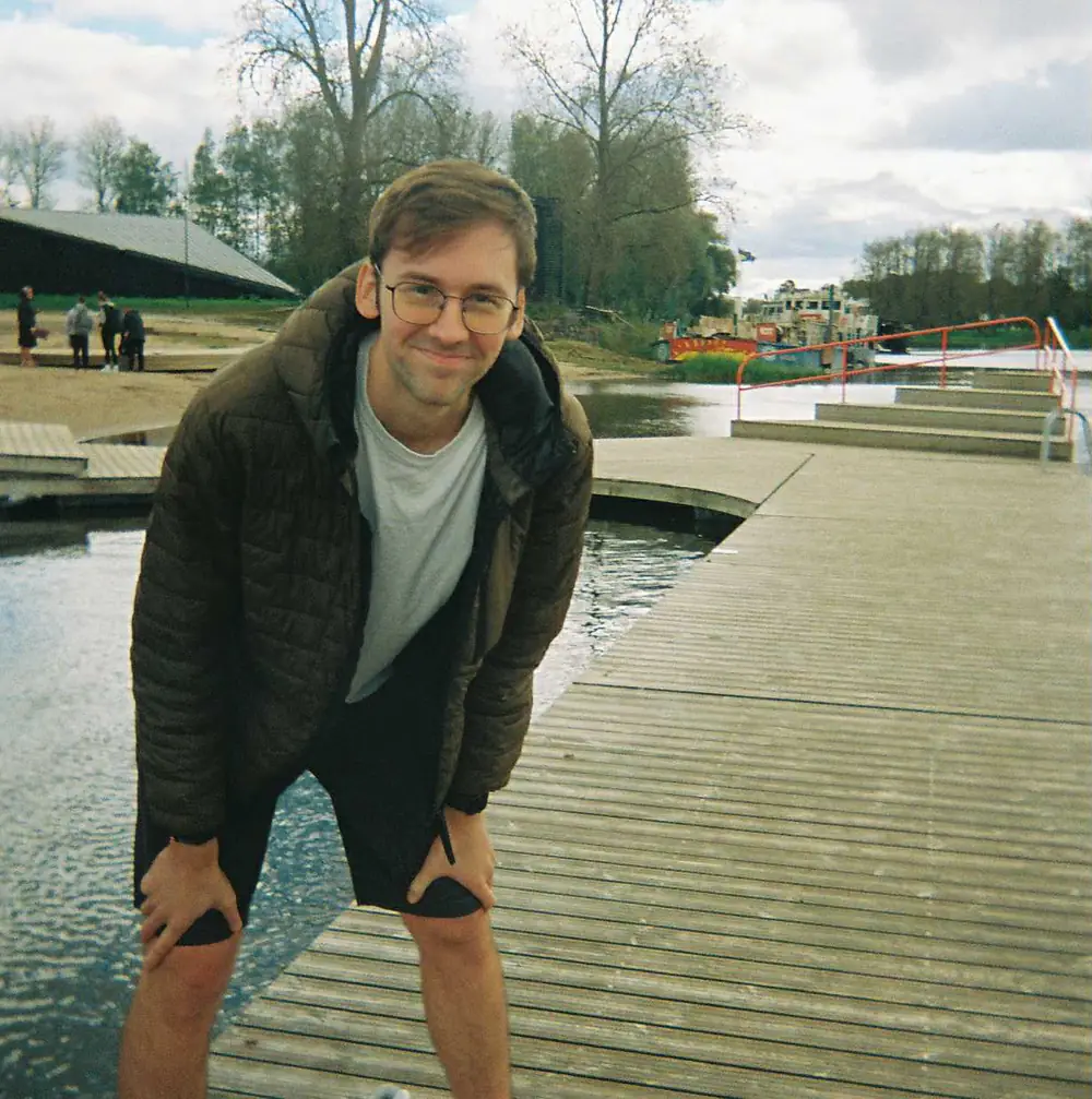 Andrius leaning forward on a wooden boardwalk by the water on a bright day